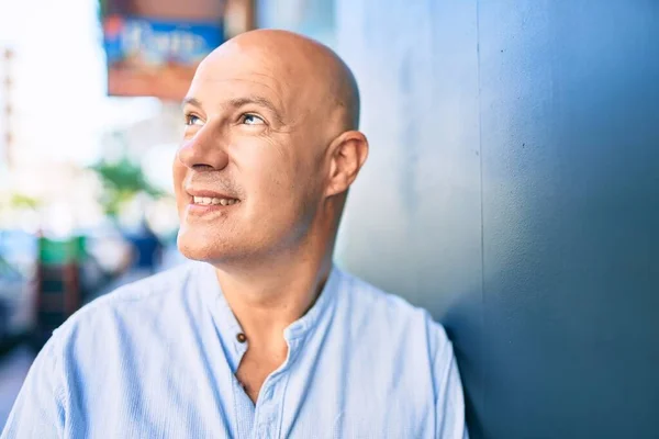 Middle age bald man smiling happy leaning on the wall at the city