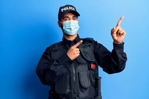 警察の制服を着た若いヒスパニック系の男と医療マスク笑顔とカメラを見て2本の手と指で側面を指して — ストック写真