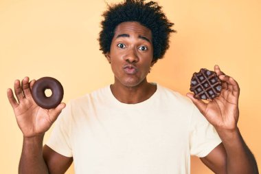 Afro saçlı, çikolatalı donut ve waffle tutan yakışıklı Afro-Amerikan erkeği öpüşürken kameraya bakıyor ve sevimli ve seksi. aşk ifadesi. 