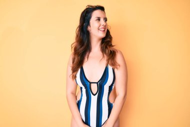 Yazlık mayo ve güneş gözlüğü takan genç, güzel esmer kadın yüzünde gülümseme, doğal bir ifade ile yan yana bakıyor. Kendinden emin gülen. 