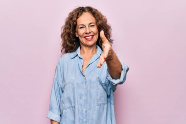 Orta yaşlı, güzel bir kadın pembe arka planda dikilmiş sıradan bir kot gömlek giyiyor. Dostça bir selamlama ve hoş geldin hediyesi olarak gülümsüyor. Başarılı bir iş..