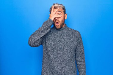 Genç, sarışın, sıradan bir süveter giyen, izole edilmiş mavi arka plan üzerinde duran, elleriyle yüzünü ve gözlerini kapatan, parmaklarından korkan genç bir adam.