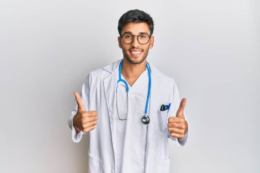 Doktor üniforması giyen genç yakışıklı bir adam ve steteskop başarı işareti. Elleriyle olumlu jest yapıyor. Gülümsüyor ve mutlu. Neşeli ifade ve kazanan jest. 