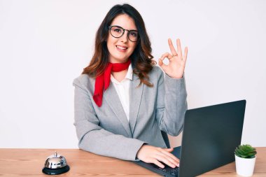 Genç güzel esmer kadın otel resepsiyonunda dizüstü bilgisayar kullanarak çalışıyor parmaklarıyla imza atıyor, gülümseyerek mükemmel bir sembol oluşturuyor. 