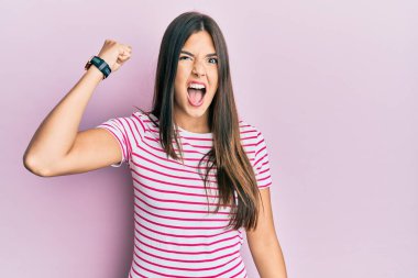 Genç esmer kadın, pembe arka plan üzerine gündelik kıyafetler giyip öfkeyle bağırırken kızgın ve kızgın bir şekilde yumruk kaldırıyor. öfke ve agresif konsept. 