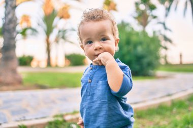 Üzgün küçük çocuk, güneşli bir günde parkta diş etlerine dokunduğu için parmaklarını dudaklarına koyuyor. Güzel sarı saçlı erkek bebek yeni bebek dişleri için acı çekiyor.