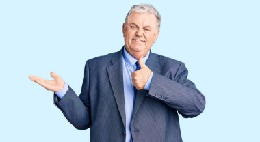 Kıdemli, gri saçlı, iş ceketi giyen, avuç içi gösteren ve baş parmağıyla hareket eden mutlu ve neşeli gülümseyen bir adam. 