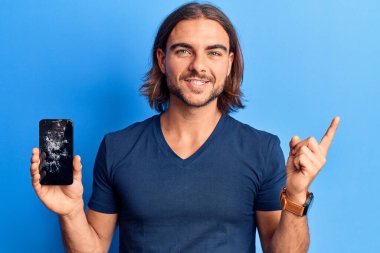 Kırık akıllı telefonu tutan genç yakışıklı adam kırık ekranı gösteriyor. Gülümsüyor, mutlu bir şekilde elini ve parmağını yana doğru gösteriyor. 