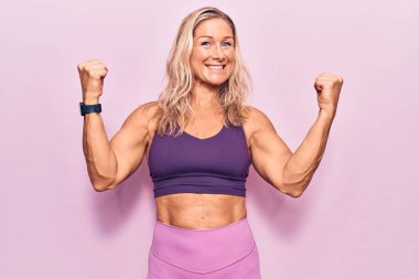 Pembe arka planda spor kıyafetleri giyen orta yaşlı sarışın kadın gururla bağırıyor, zaferi kutluyor ve kollarını kaldırarak başarıyı kutluyor. 