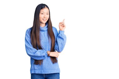 Genç, güzel, balıkçı yaka kazak giyen, yüzünde kocaman bir gülümseme olan, eli ve parmağı kameraya bakan Çinli bir kadın.. 