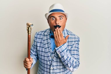 Bıyıklı, klasik ve zarif moda tarzıyla ağzı elle kaplanmış, şok olmuş ve hata yapmaktan korkan olgun bir Orta Doğulu. şaşırmış ifade 