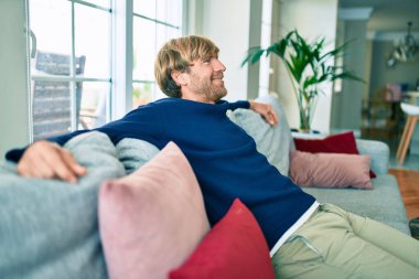 Orta yaşlı, yakışıklı, beyaz bir adam evinde kanepede oturuyor.
