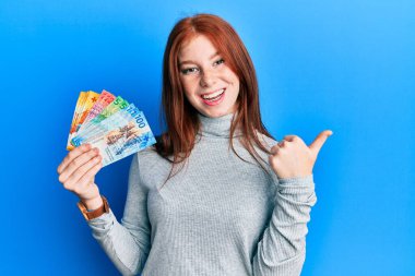 Genç kızıl saçlı kız İsviçre frangı banknotları tutuyor başparmağıyla yan tarafa bakıyor mutlu bir şekilde gülümsüyor. 