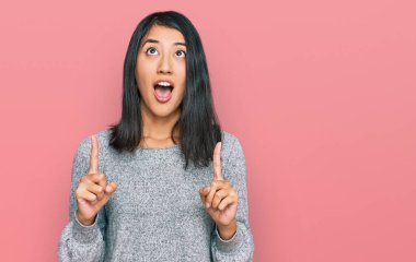 Güzel Asyalı genç kadın gündelik kıyafetler giyip şaşkın şaşkın yukarı bakıyor ve parmaklarıyla işaret ediyor ve kollarını kaldırıyor.. 