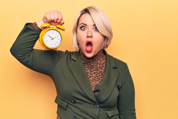 Young beautiful plus size blonde woman holding vintage alarm clock over yellow background scared and amazed with open mouth for surprise, disbelief face