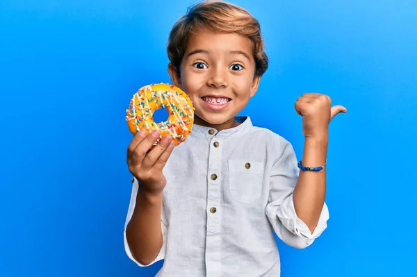 Adorable Niño Latino Sosteniendo Donut Apuntando Pulgar Hacia Lado Sonriendo — Foto de Stock
