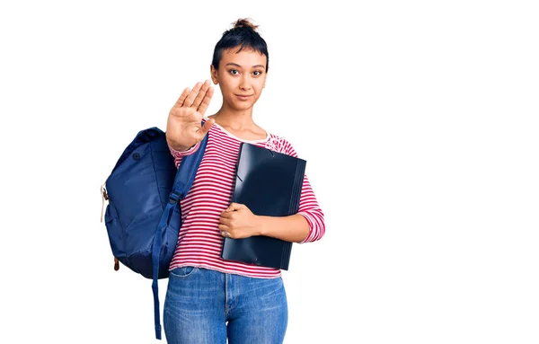 年轻女子背着学生背包 张开手拿着活页夹 做了个手势 脸上带着严肃而自信的表情和防守姿势 — 图库照片