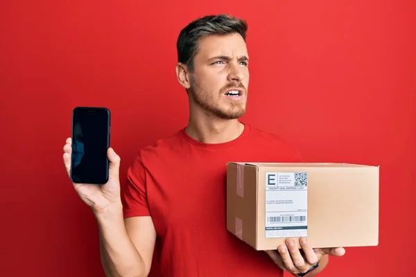 Handsome Caucasian Man Holding Delivery Package Showing Smartphone Screen Angry — Stock Photo, Image