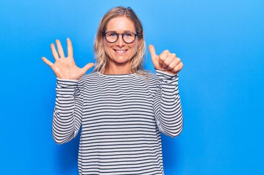 Orta yaşlı, beyaz, çizgili süveter giyen ve gözlük takan sarışın kadın altı numaralı parmaklarını gösterip gösterirken kendinden emin ve mutlu bir şekilde gülümsüyor.. 