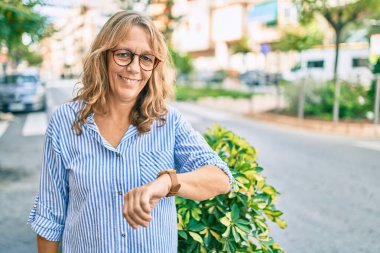 Orta yaşlı, beyaz, gülümseyen bir kadın. Mutlu görünümlü bir saat şehre bakıyor..