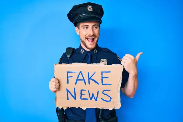 Young caucasian man wearing police uniform holding fake news banner pointing thumb up to the side smiling happy with open mouth