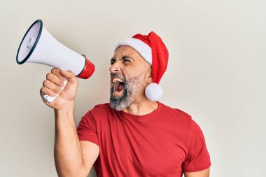 Orta yaşlı, gri saçlı, Noel Baba şapkalı bir adam izole edilmiş beyaz arka plan üzerinde megafon kullanarak bağırıyor..
