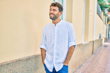 Güneşli bir günde günlük beyaz gömlek giyen sakallı yakışıklı adam dışarıda mutlu bir şekilde gülümsüyor.