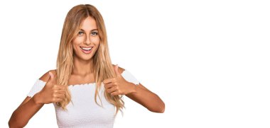 Güzel sarışın genç bir kadın, günlük beyaz tişört giyer, başarı işareti yapar, elleriyle olumlu jest yapar, gülümser ve mutlu olur. Neşeli ifade ve kazanan jest. 