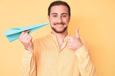 Elinde kağıt uçak tutan yakışıklı adam mutlu ve pozitif gülümsüyor, başparmağıyla mükemmel ve onay işareti yapıyor. 