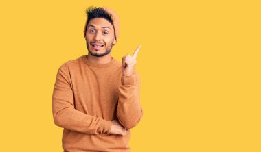 Yakışıklı Latin Amerikalı genç adam, kışlık süveter ve yüzünde kocaman bir gülümseme olan yün şapkalı, eli ve parmağı kameraya bakıyor.. 