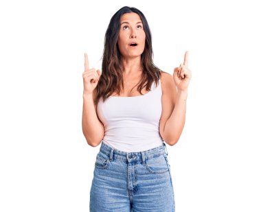 Kolsuz tişört giyen genç ve güzel esmer kadın şaşırmış ve şaşırmış bir şekilde yukarı bakıyor ve parmaklarıyla işaret ediyor ve kollarını kaldırıyor.. 