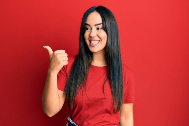 Gündelik kırmızı tişört giyen genç ve güzel bir İspanyol kız başparmağıyla yan tarafa bakıyor ve ağzı açık gülümsüyor. 