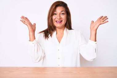 Orta yaşlı esmer, Latin iş kadını, gündelik beyaz gömlekli, çılgın bir şekilde masada oturuyor ve kollarını kaldırıp heyecanla haykırarak başarıyı kutluyor. Kazanan konsept 