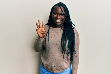Saçları örgülü, günlük kıyafetler giyen ve gözlüklü genç siyahi bir kadın üç numarayı gösterip işaret ederken kendinden emin ve mutlu bir şekilde gülümsüyor.. 