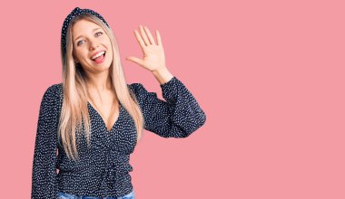 Genç ve güzel sarışın kadın gündelik giysiler içinde merhaba diyor mutlu ve gülümsüyor, dostça karşılama jesti yapıyor. 