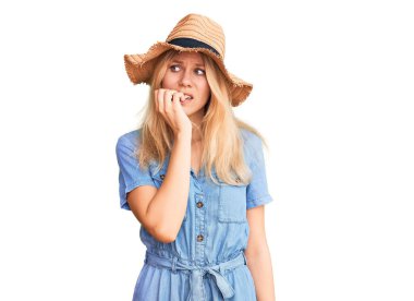 Yazlık şapkası ve elbisesi olan genç ve güzel sarışın kadın gergin görünüyor ve elleri ağızdan tırnak yiyor. anksiyete. 