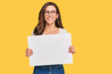 Boş pankart tutan genç esmer kadın pozitif ve mutlu görünüyor ve kendine güvenen bir gülümsemeyle dişlerini gösteriyor. 