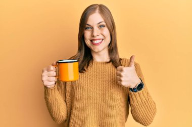 Güzel beyaz kadın kahve içiyor mutlu ve pozitif gülümsüyor, başparmağıyla mükemmel ve onay işareti yapıyor. 