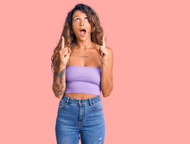 Gündelik giysiler giyen Latin kökenli genç bir kadın şaşırmış ve şaşırmış bir şekilde yukarı baktı ve parmaklarıyla işaret ederek kollarını kaldırdı.. 