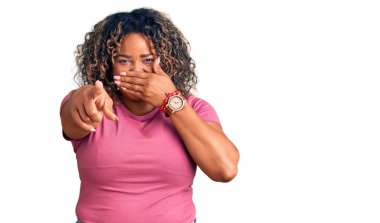 Genç Afro-Amerikan artı cüsseli kadın sana gülüyor, eli ağzında kamerayı işaret ediyor, utanç ifadesi var. 