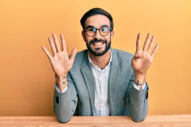 Ofiste çalışan Latin asıllı genç adam dokuz numaralı parmaklarını gösterip işaret ederken kendinden emin ve mutlu bir şekilde gülümsüyor.. 