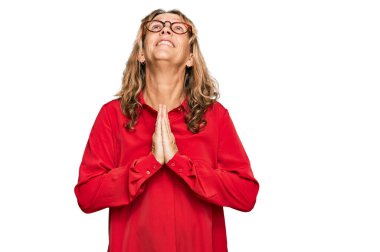 Orta yaşlı sarışın kadın kırmızı arka plan üzerine gündelik tişört giyip elleri ile yalvarıyor ve dua ediyor. Yüzünde umut ifadesi var. Çok duygusal ve endişeli. Yalvarıyor. 