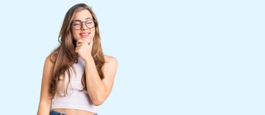 Gündelik kıyafetler giyen, çenesinde elleri olan, soru soran, düşünceli, genç, güzel beyaz bir kadın. Düşünceli bir yüzle gülümsüyor. şüphe kavramı. 