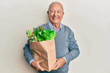 Elinde ekmek ve bakkaliye poşeti olan beyaz bir adam pozitif ve mutlu görünüyor ve kendine güvenen bir gülümsemeyle dişlerini gösteriyor. 