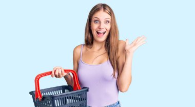 Genç, sarışın bir kadın elinde alışveriş sepeti tutarak zaferi mutlu bir gülümsemeyle ve ellerini kaldırarak kutluyor. 