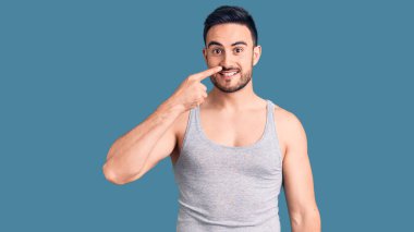 Mayolu ve kolsuz tişörtlü genç ve yakışıklı bir adam. Parmakla yüzünü ve burnunu gösteriyor. Neşeli bir şekilde gülümsüyor. güzellik konsepti 