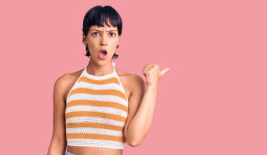 Kısa saçlı esmer bir kadın, sıradan kıyafetler giyiyor. El parmağı yana dönük, ağzı açık, şaşkın bir şekilde şaşırmış.. 