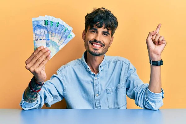 Joven Hispano Sosteniendo Billetes Rand Sudafricanos Sentado Mesa Sonriendo Feliz —  Fotos de Stock