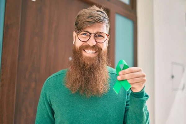 Young irish man with redhead beard smiling happy holding green awareness ribbon at the city.
