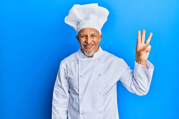 自信と幸せ笑顔ながら プロの料理の制服と帽子を身に着けている中世の白髪の男は指の数3で現れ 指摘します — ストック写真